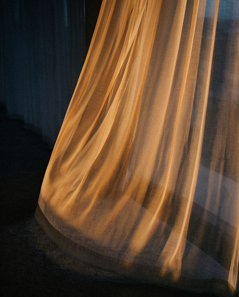 A heavy fabric curtain half drawn on a rod, seen from a low off-axis angle under warm side light against a dark ground, caught mid-pull with the folds in motion