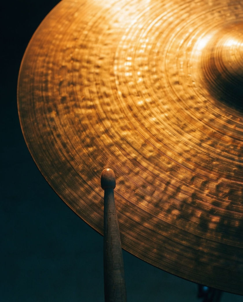 A bronze crash cymbal struck by a stick in warm low light against a dark ground, metal shimmering at the point of impact