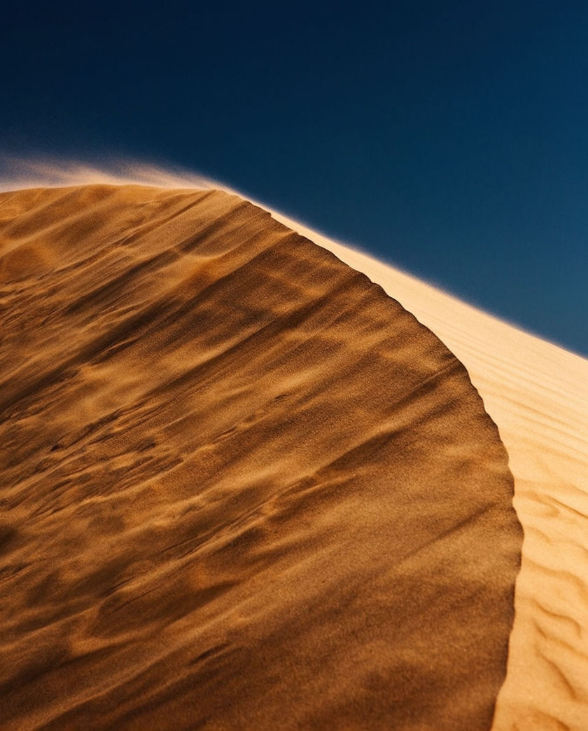 A single dune crest with a plume of sand streaming off its ridge under warm low light against a dark sky, grit lifting where the wind catches it