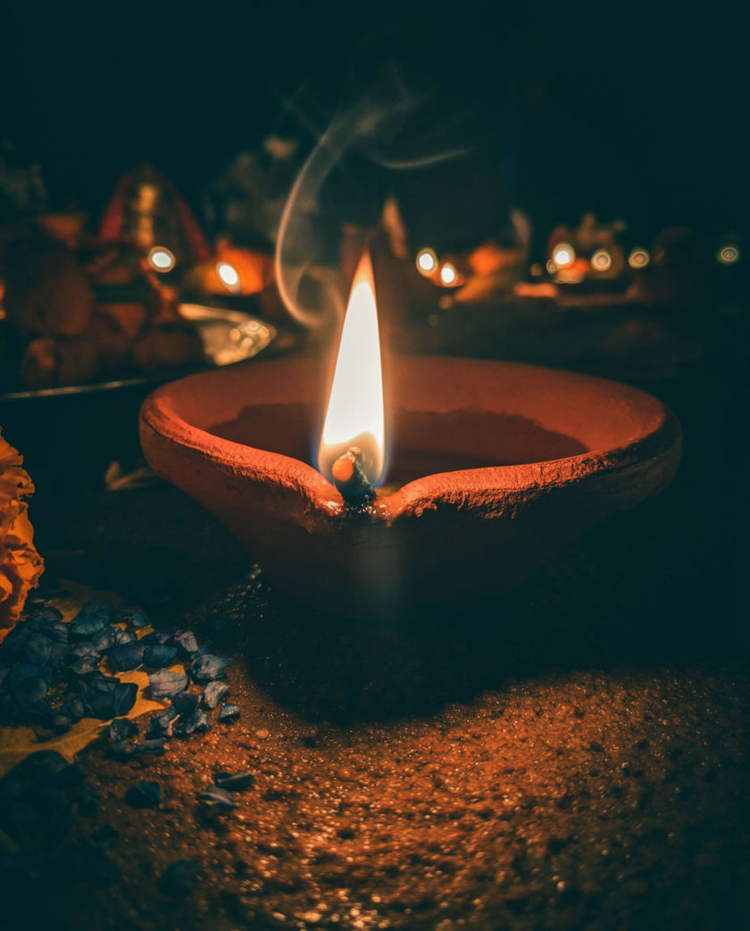 A row of glowing clay diya lamps with a sparkler beside them under one warm low light against a dark ground, sparks flying