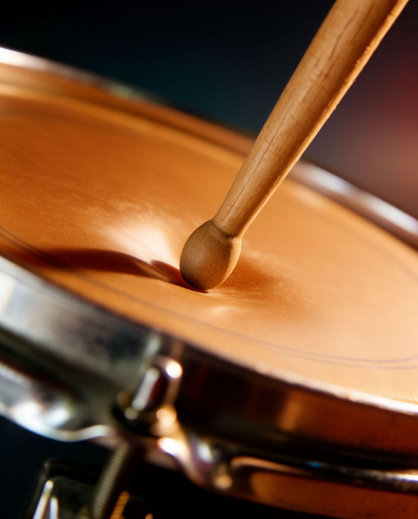 A wooden drum stick striking a snare drumhead in warm low light against a dark ground, skin flexing at impact
