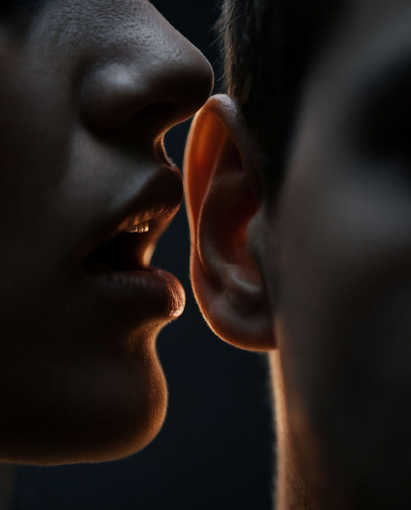 A shadowed mouth close to an ear lit by one warm rim of light against a cold black ground, lips parted mid-whisper