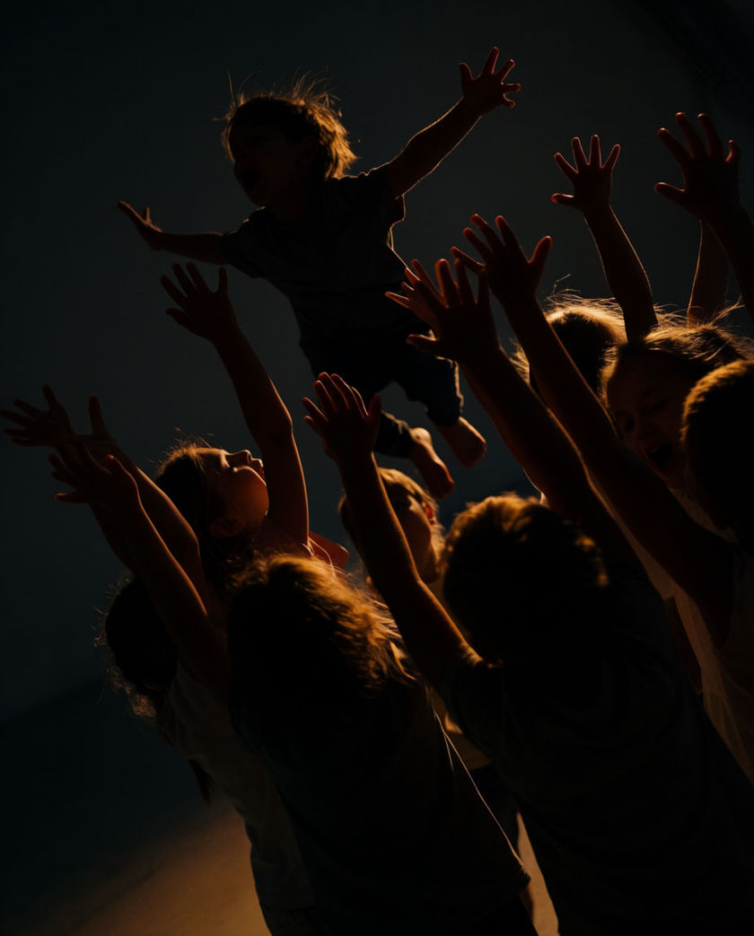 A small child with arms thrown up mid-cheer, warm side light catching the raised hands against a dark ground, mouth open in a shout