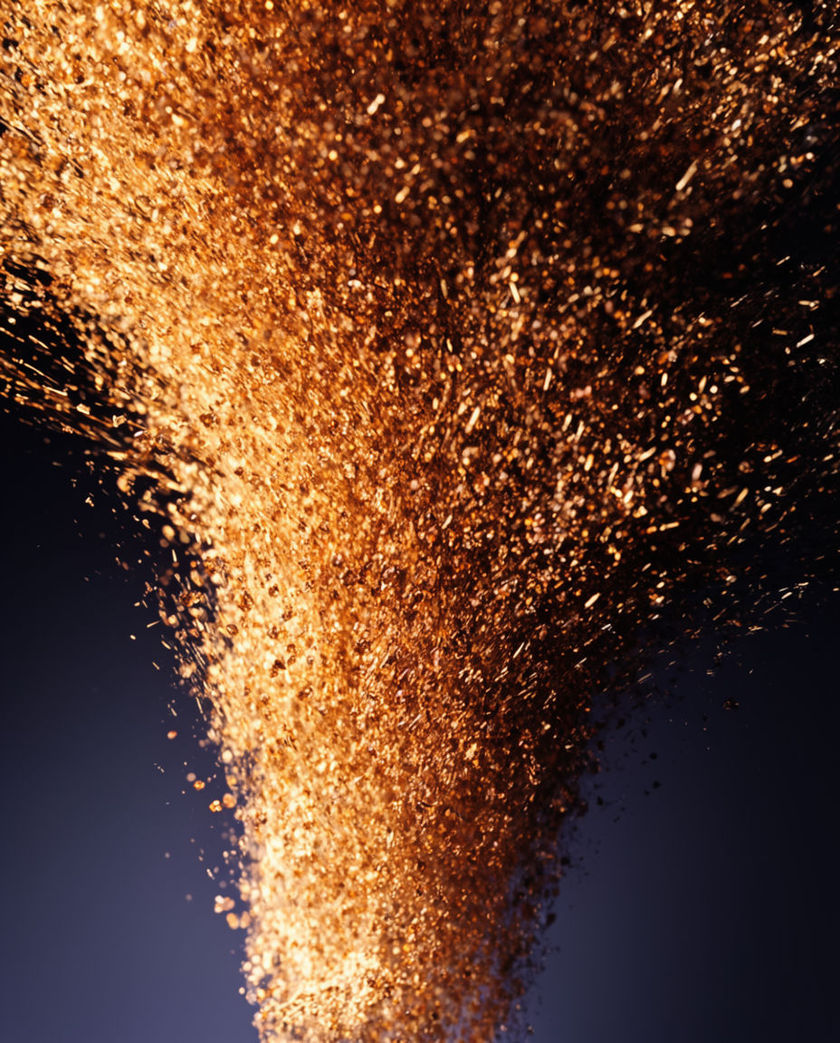 A rising column of golden light and sparks bursting upward out of a dark surface under one warm side light, seen from a low angle