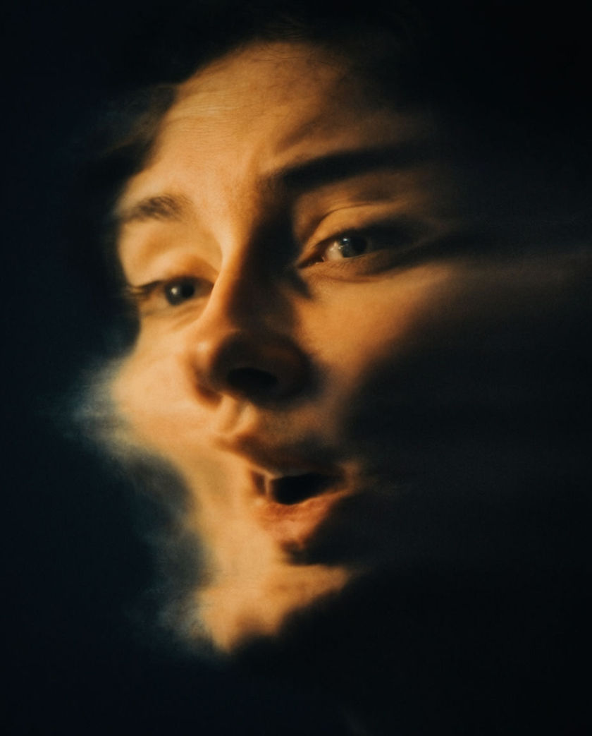 A warped distorted face dissolving into shadow under warm low light against a cold black ground, edges smearing
