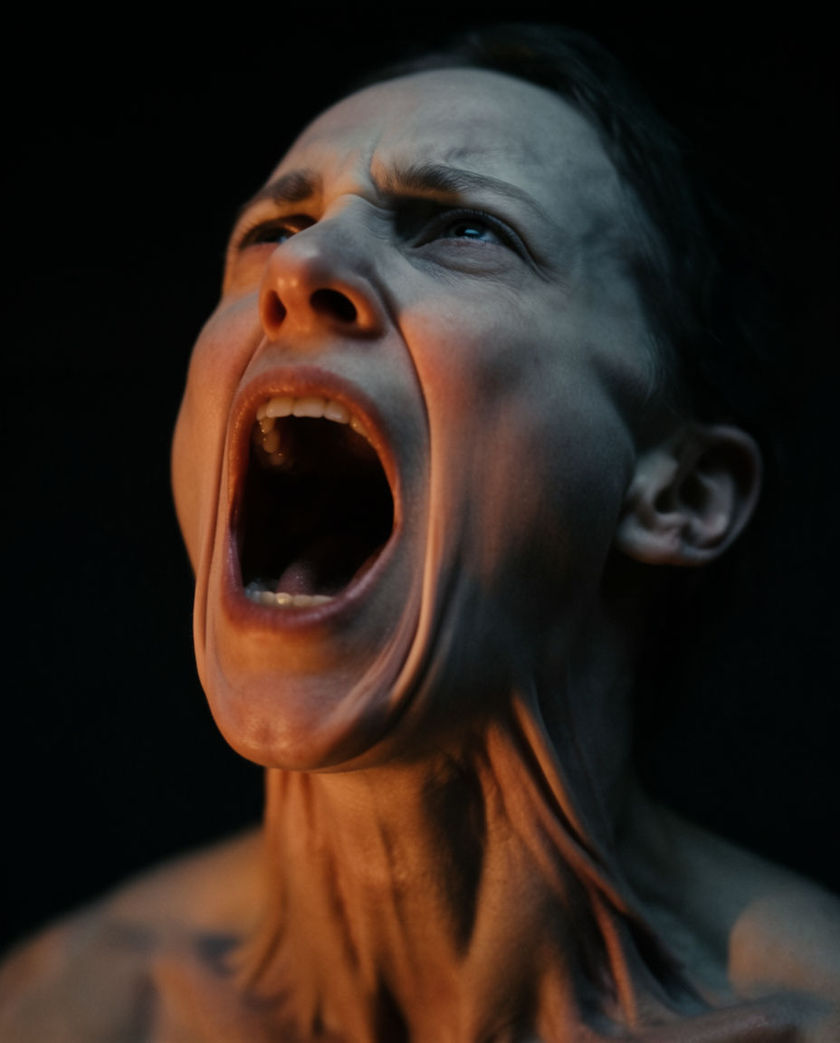 A pale contorted face tilted back under warm low light against a cold black ground, mouth stretched mid-scream