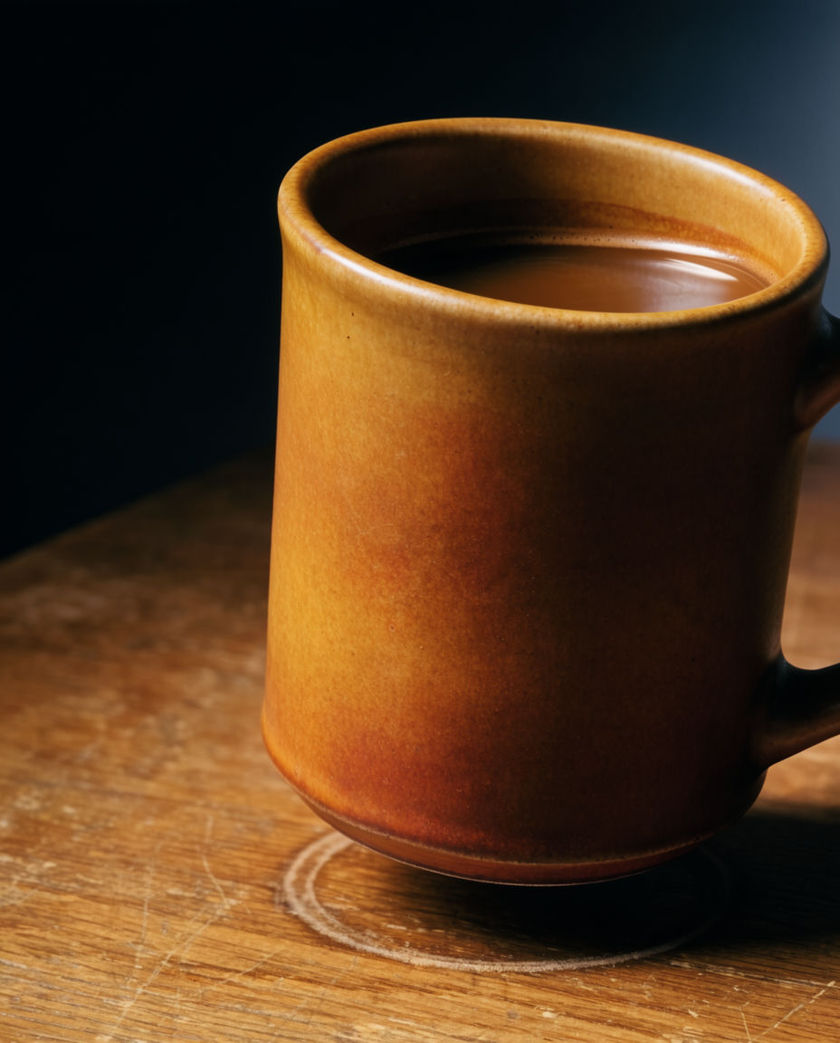 A hand setting a ceramic mug on a wooden table, filling the frame from a low off-axis angle with one warm directional light against a dark ground