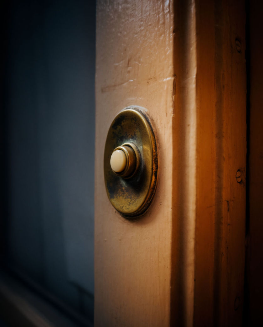 A thumb pressing a round lit doorbell button on a doorframe under warm low side light against a dark ground, button caught mid-press