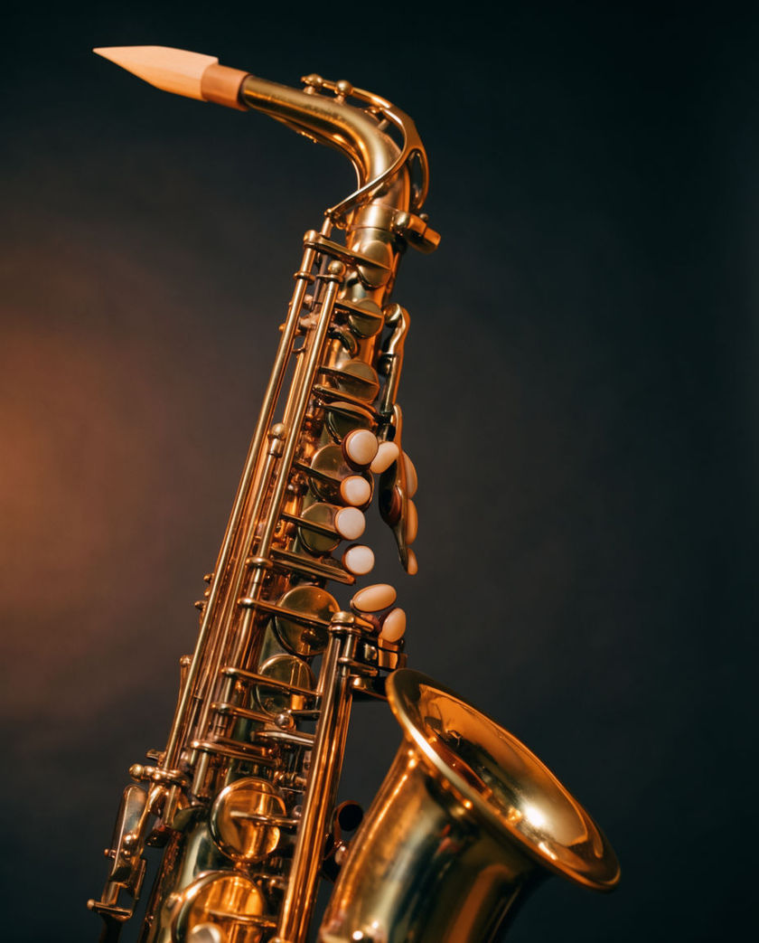 The brass keys and bell of a saxophone catching warm low light against a dark ground, a hand poised on the keys mid-note