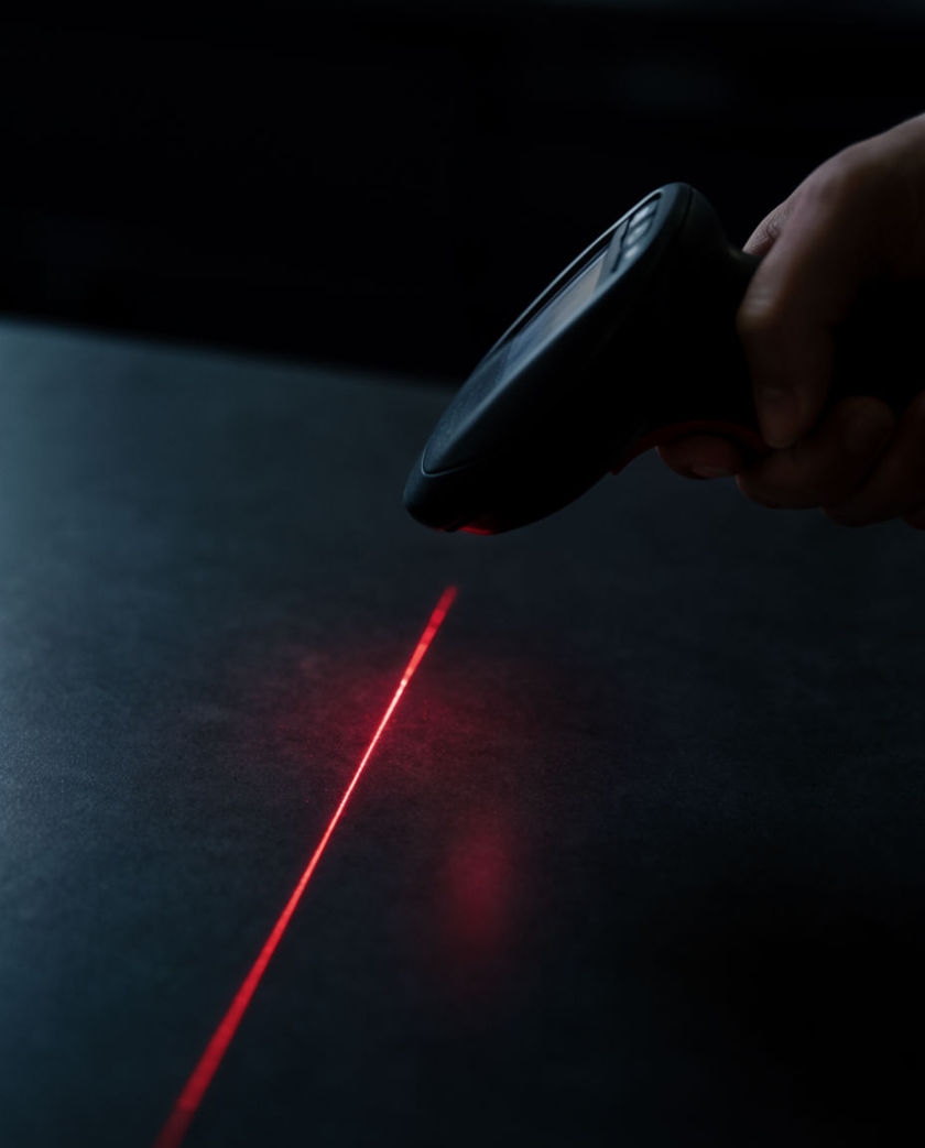 A handheld barcode scanner throwing a thin red laser line across a dark surface under one warm side light, seen from a low off-axis angle
