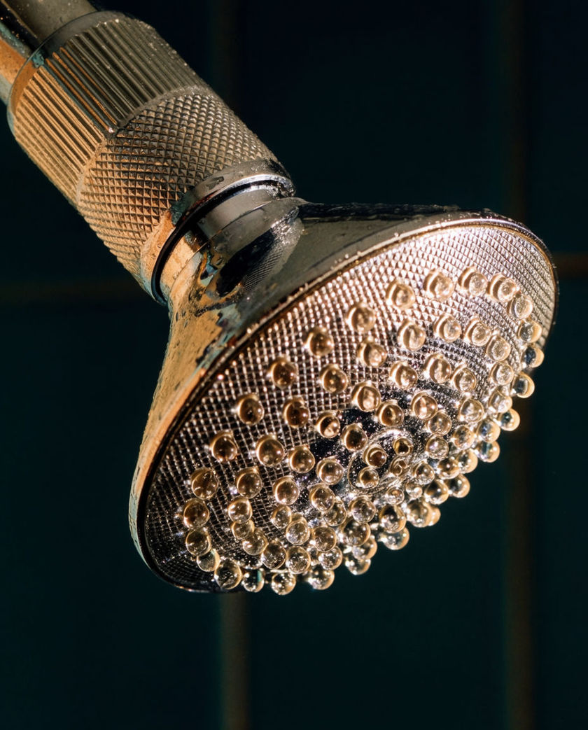 A chrome showerhead streaming water caught mid-spray under warm low side light against a dark ground, droplets lit as they fall