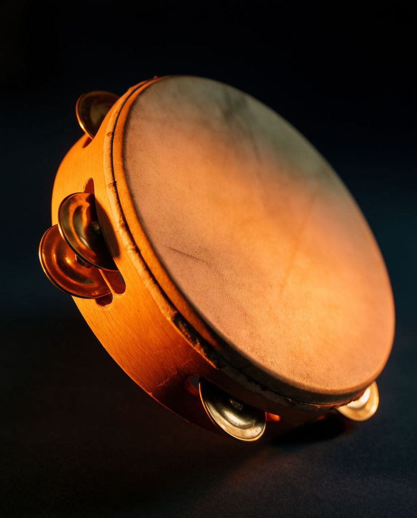 A hand-held tambourine caught mid-strike under one warm side light against a dark ground, zils blurred as they ring