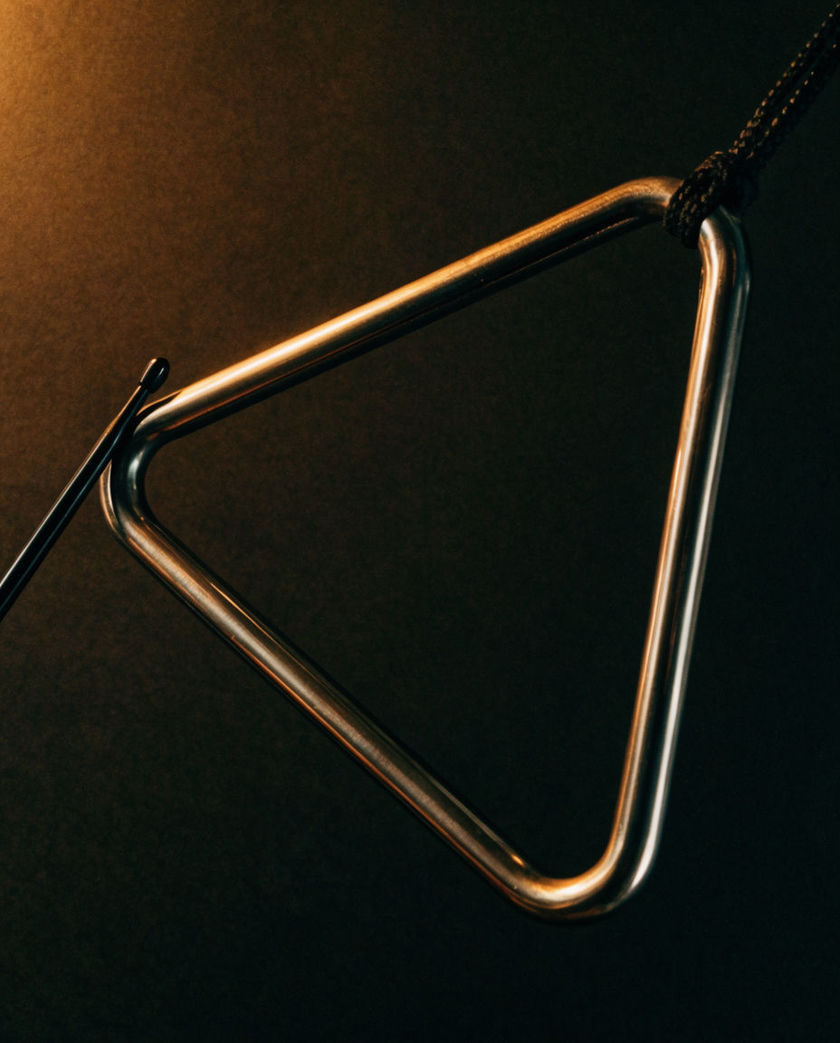 A steel percussion triangle hanging from a cord, struck by a beater under one warm side light against a dark ground, ringing