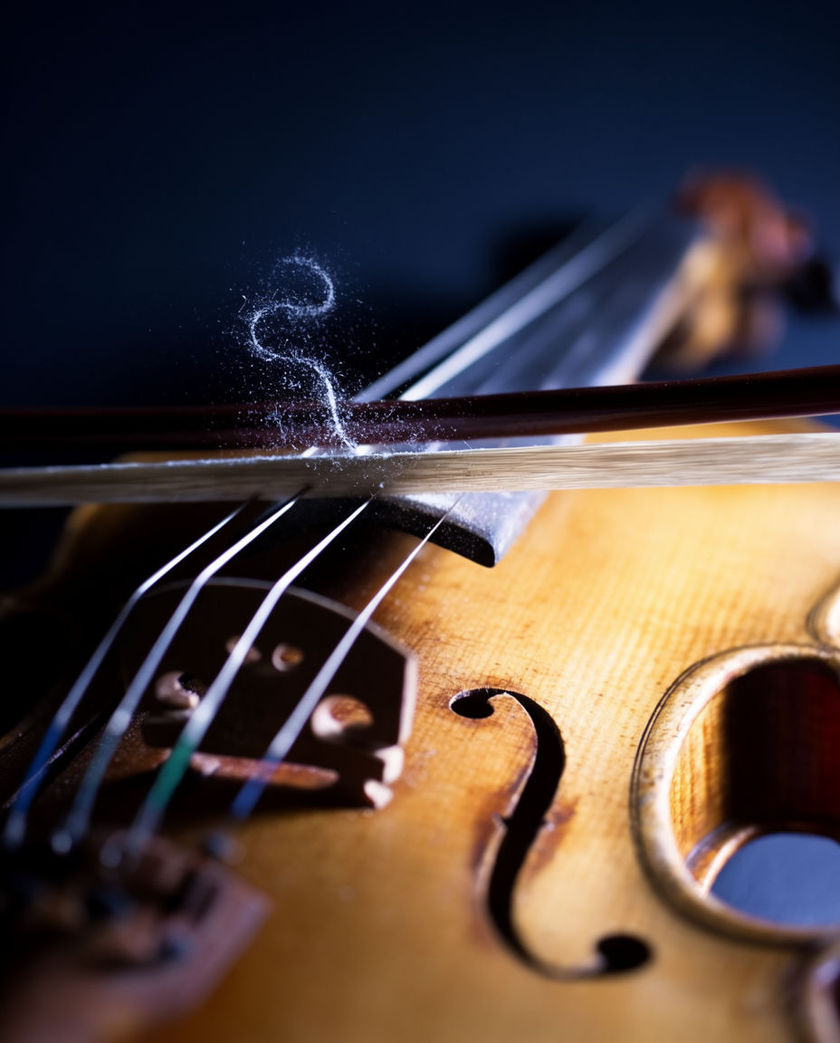 A bow drawn across violin strings in warm low light against a dark ground, horsehair pressing into a string mid-note