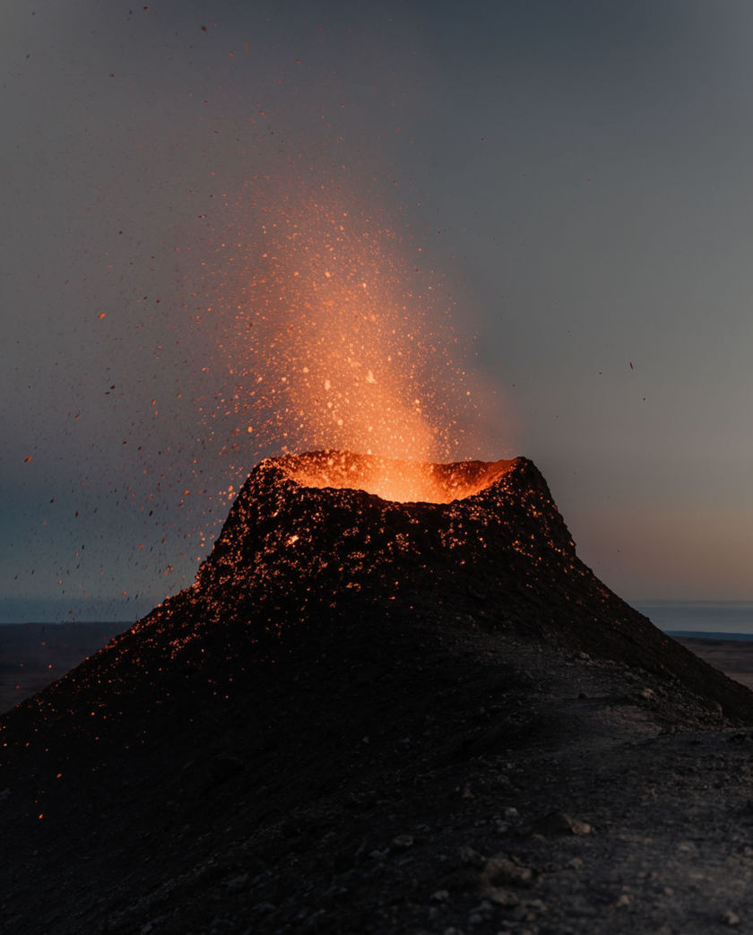 A volcanic crater seen from a low angle throwing a plume of glowing lava and sparks into a dark sky, molten light raking the rock