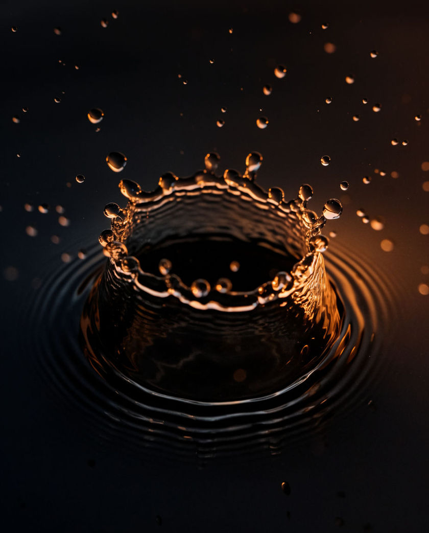 A single water drop falling toward a dark still basin under one warm side light, ripples spreading from the impact