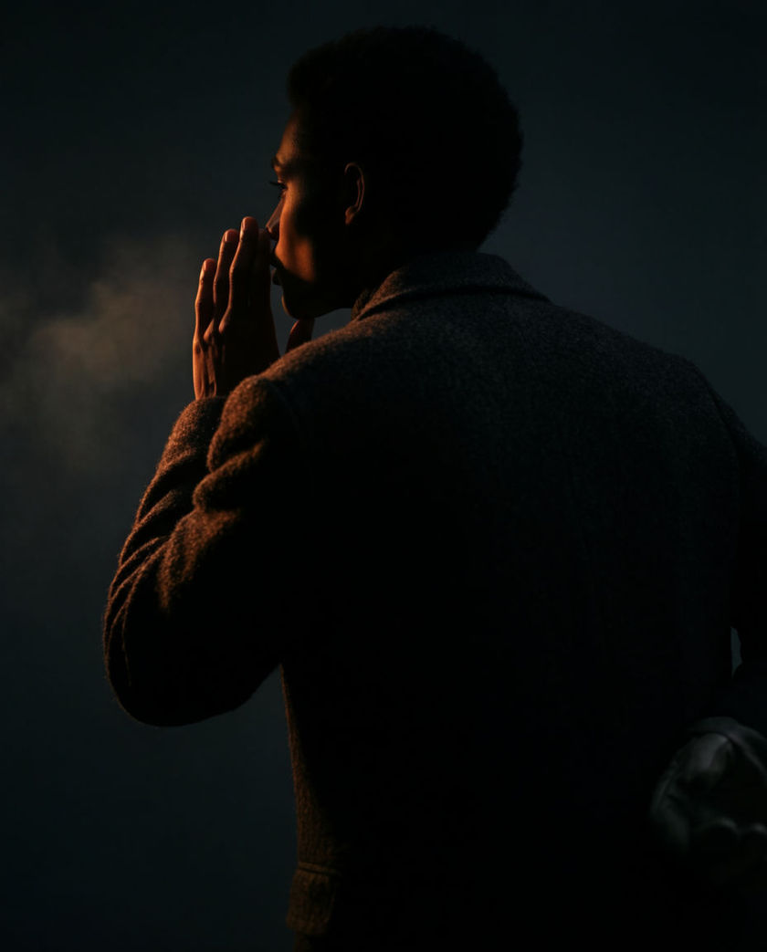 A cupped hand raised close to slightly parted lips seen from the side, one warm light grazing the mouth against a deep dark ground, mid-whisper