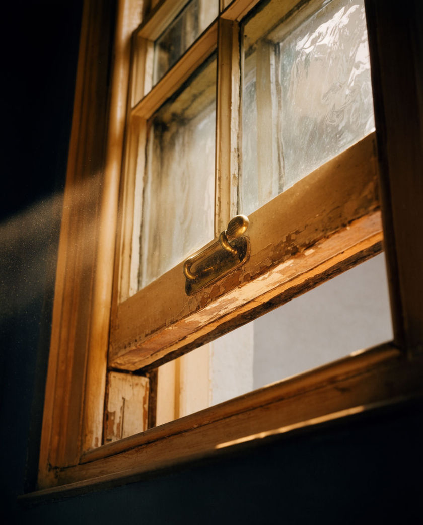 A wooden sash window pushed halfway up, seen from a low off-axis angle under warm side light against a dark ground, caught mid-slide