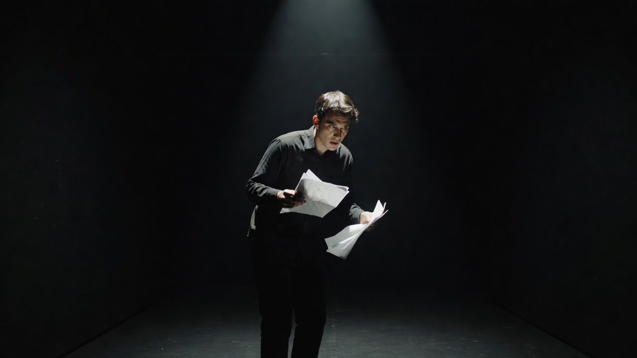 Solo performer reading from loose pages under a single spotlight in a black-box space
