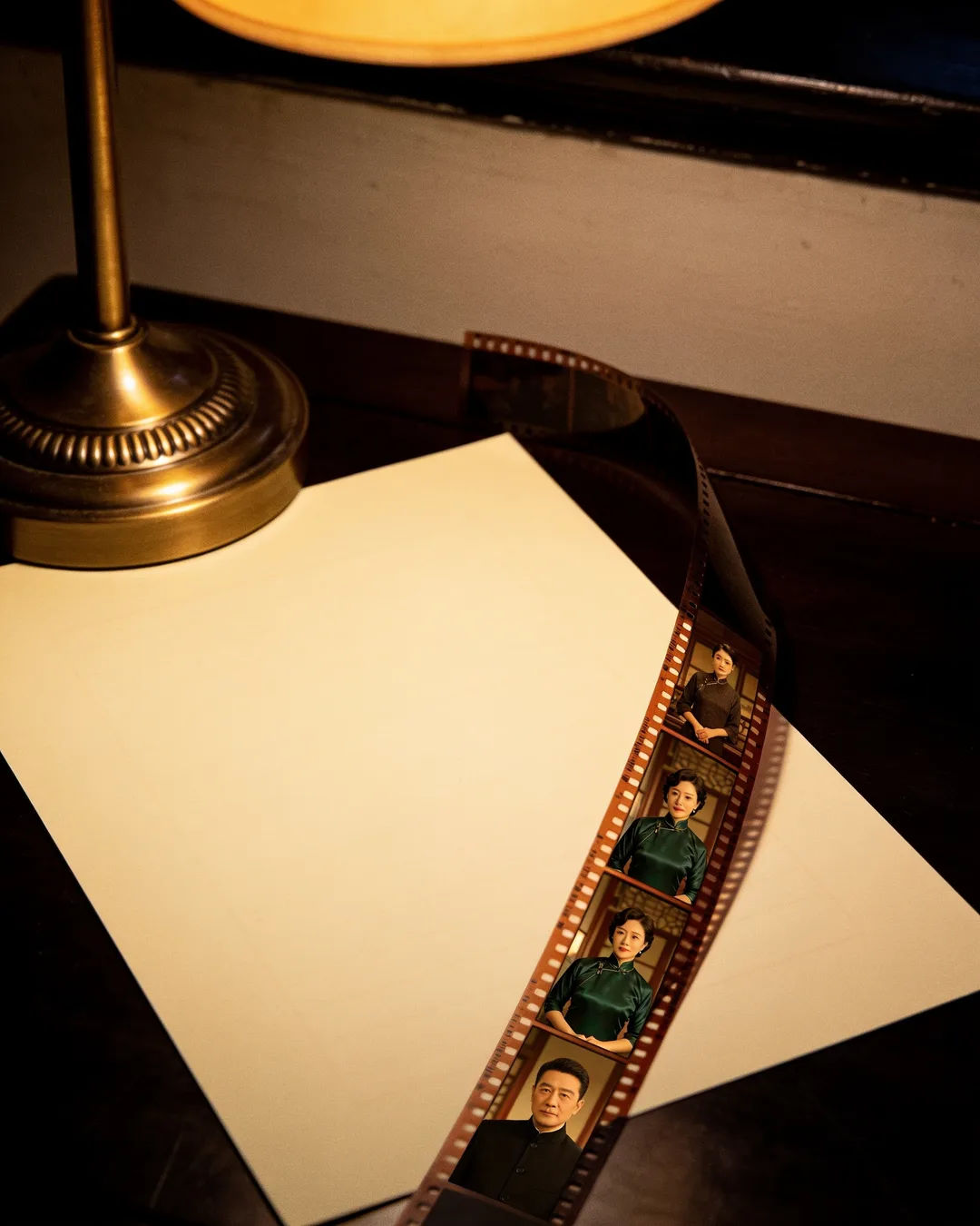 A blank page under a desk lamp beside a strip of film frames from a Chinese family drama