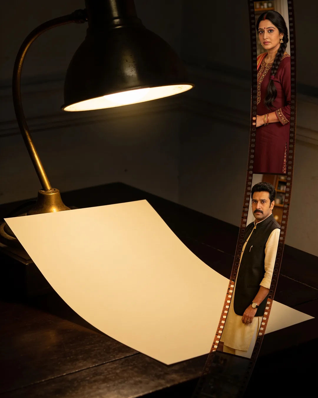 A blank page under a desk lamp beside a strip of film frames from a Hindi family drama