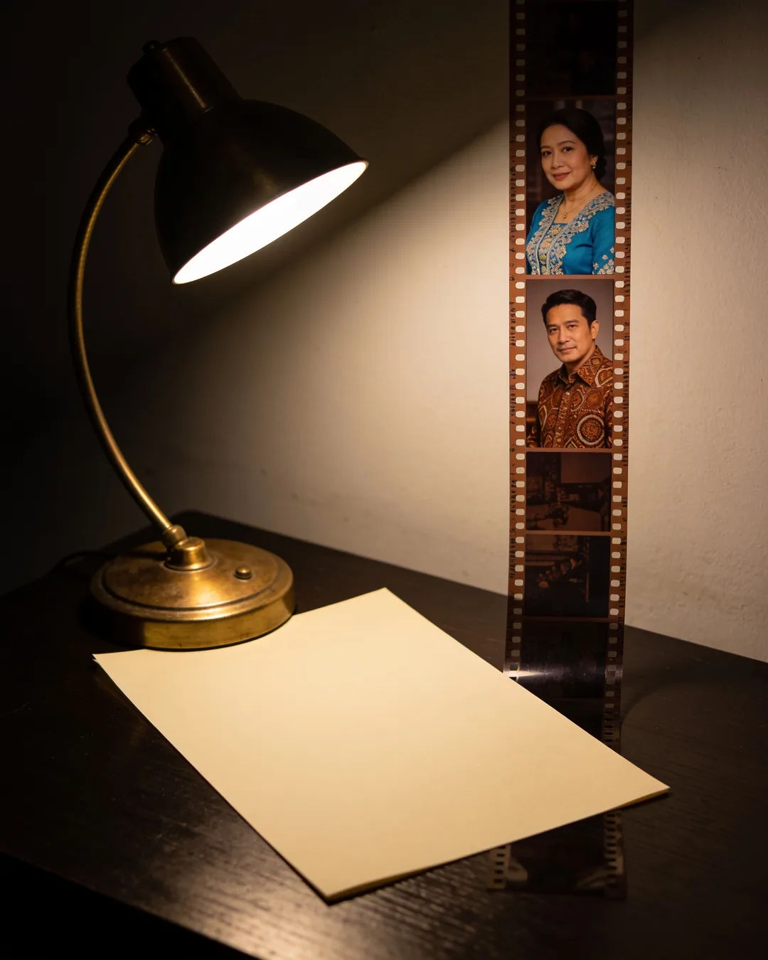 A blank page under a desk lamp beside a strip of film frames from an Indonesian family drama