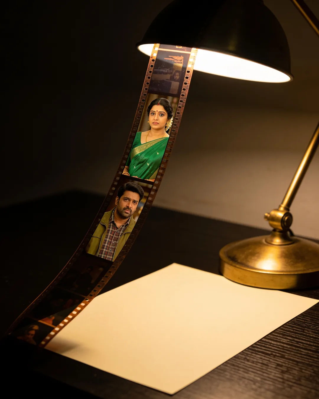 A blank page under a desk lamp beside a strip of film frames from a Kannada family drama
