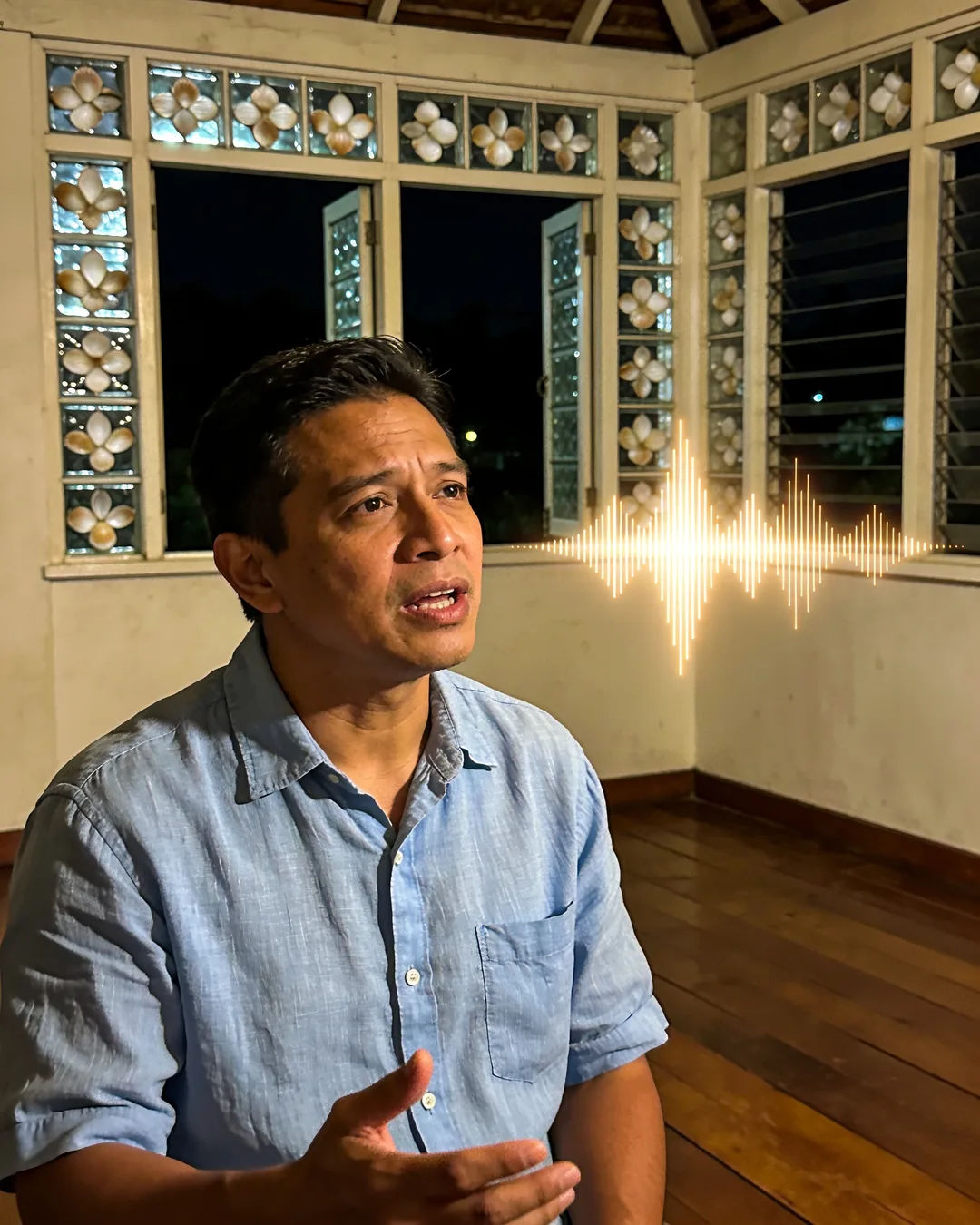 A Filipino man in a blue linen shirt speaking at home, a sound waveform beside him