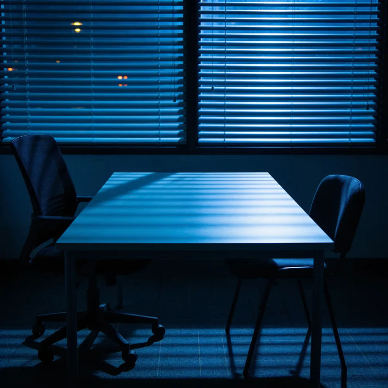 A dark office with two chairs facing each other across a bare desk in striped blue light