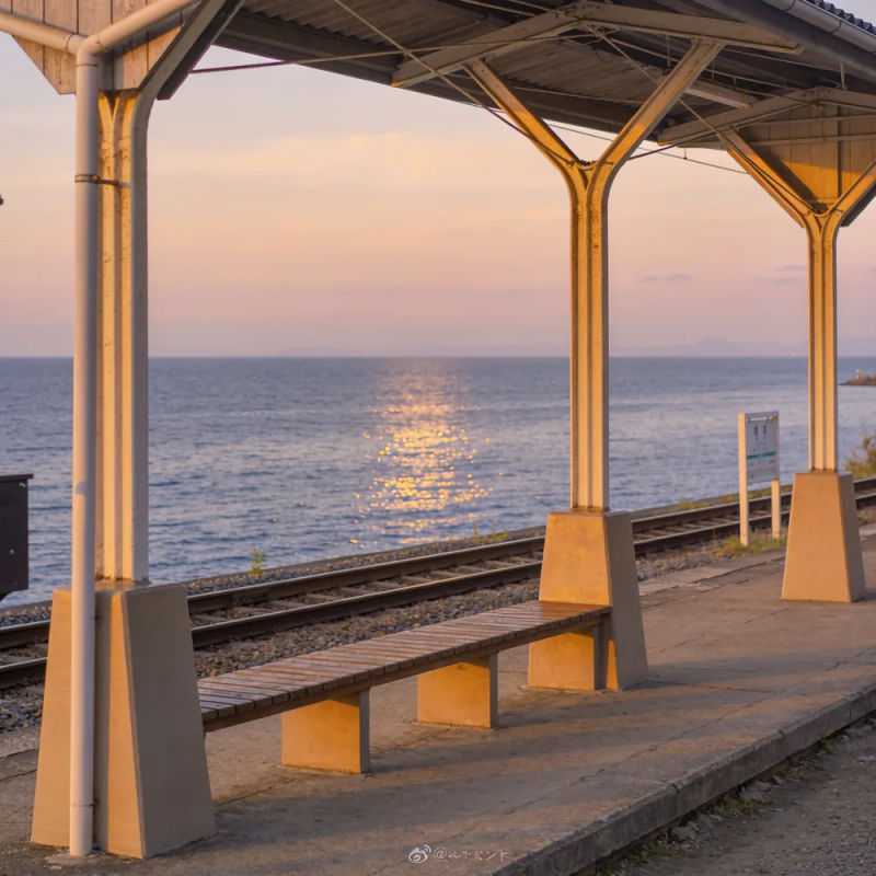 A tiny seaside train platform at dusk with the sea glinting beyond the tracks