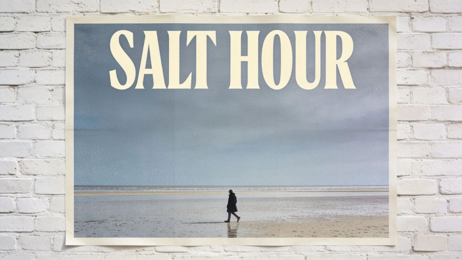 A printed poster on a whitewashed brick wall, the title SALT HOUR in tall cream letters above a lone figure walking a shoreline