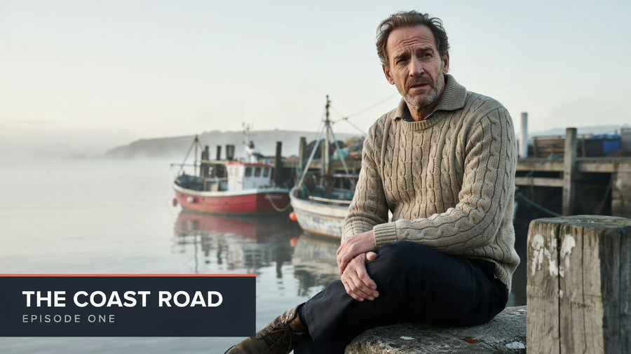 An interview frame of a fisherman by a misty harbor with a lower-third reading THE COAST ROAD