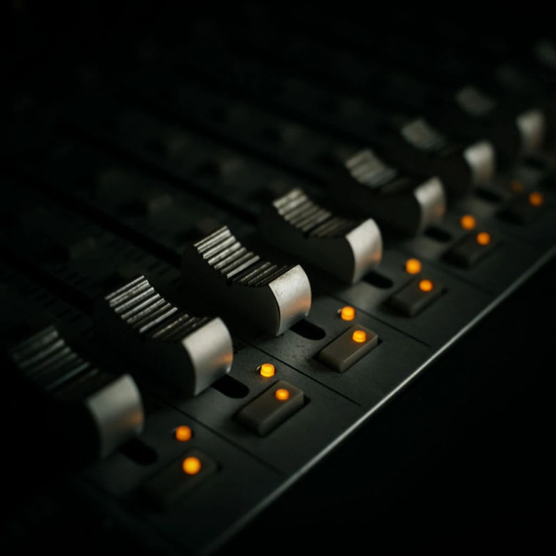 A row of studio mixer faders set at descending heights, amber indicator lights glowing