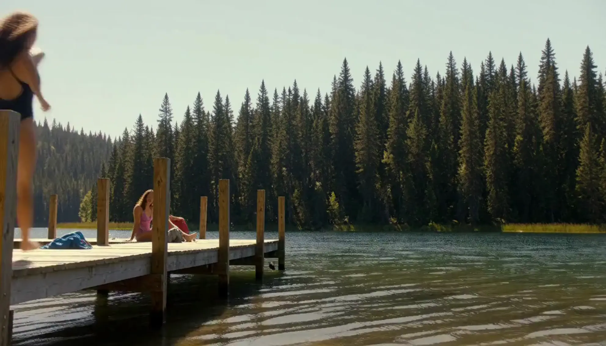 A young woman diving off a wooden dock into a lake ringed by pine forest