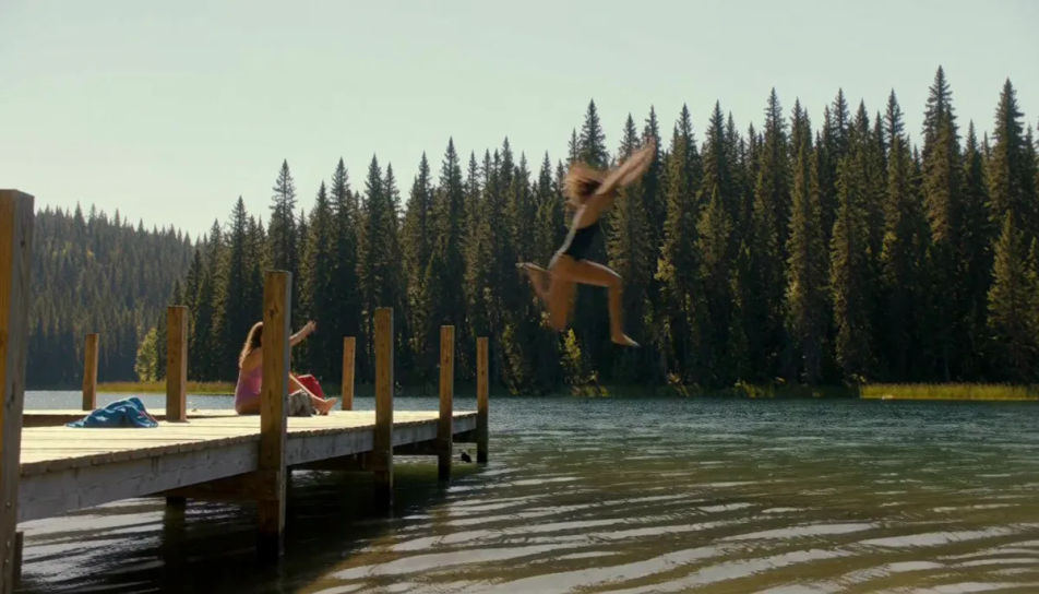 A young woman diving off a wooden dock into a lake ringed by pine forest