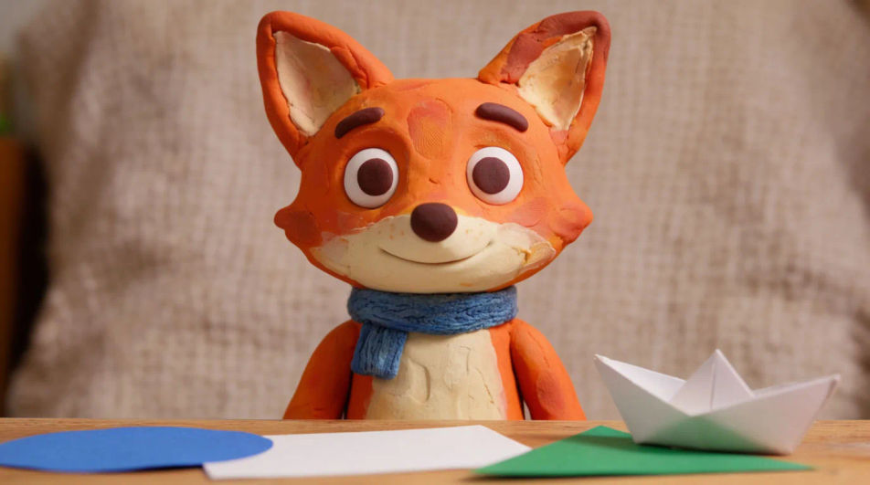 A clay fox character in a blue scarf sitting at a craft table with a paper boat