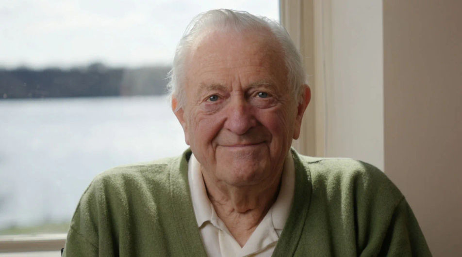 An elderly man in a green cardigan by a window overlooking a winter river