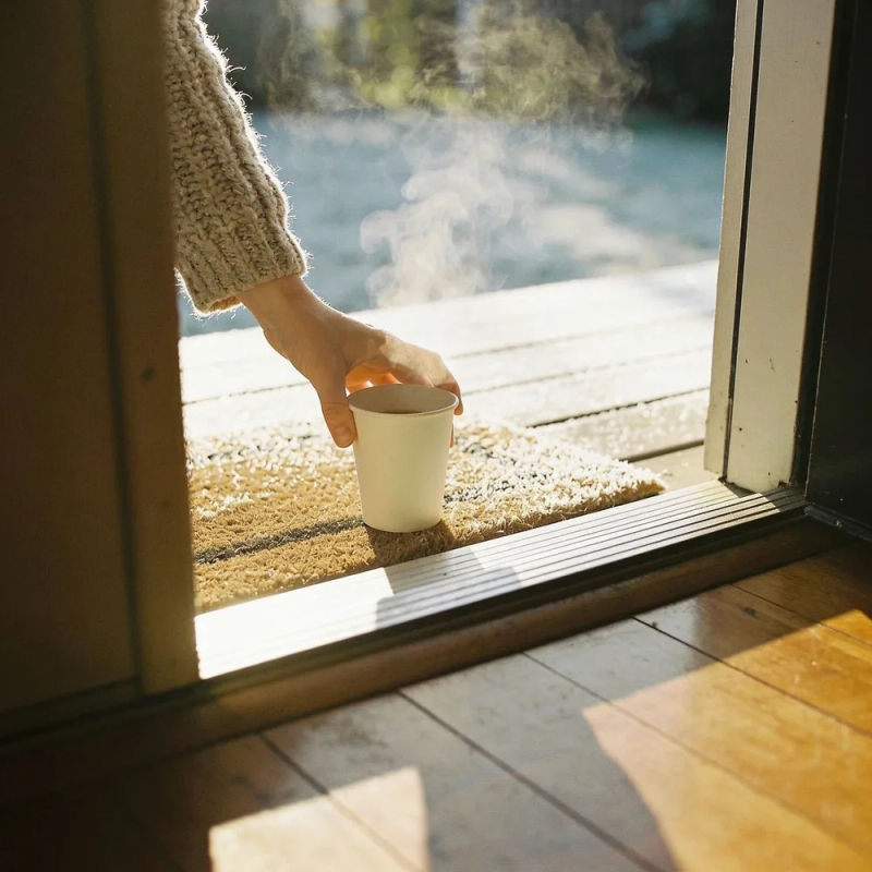 A hand reaching through a front door for a steaming paper cup left on the doormat