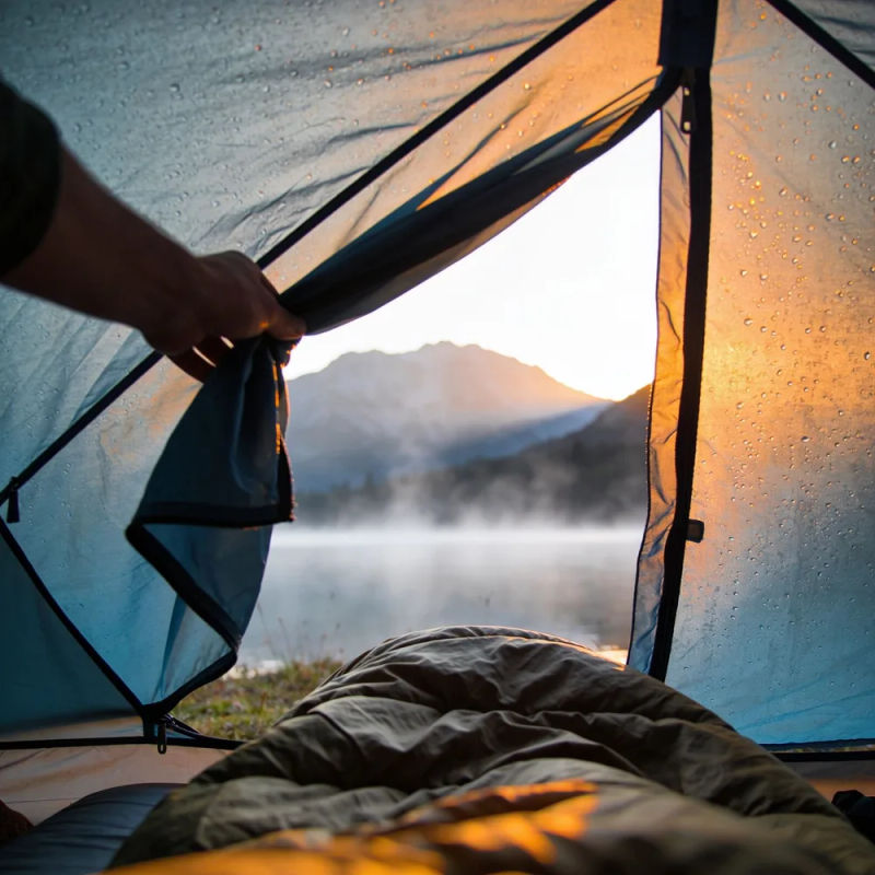 A hand unzipping a tent door at first light onto a misty mountain lake