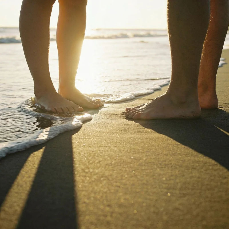 Two pairs of bare feet in wet sand as a wave pulls back at sunset