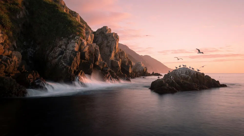 Rugged coastline with sea cliffs and crashing waves at dawn under a soft pink sky