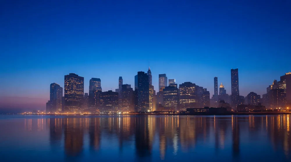 City skyline against the horizon at blue-hour dusk with lit windows and a river reflection