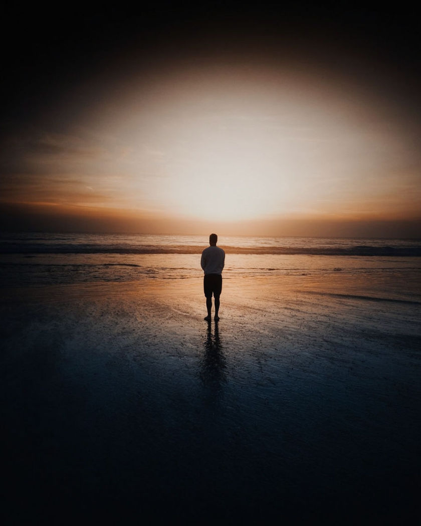 A figure seen from behind standing on wet sand facing a golden ocean horizon