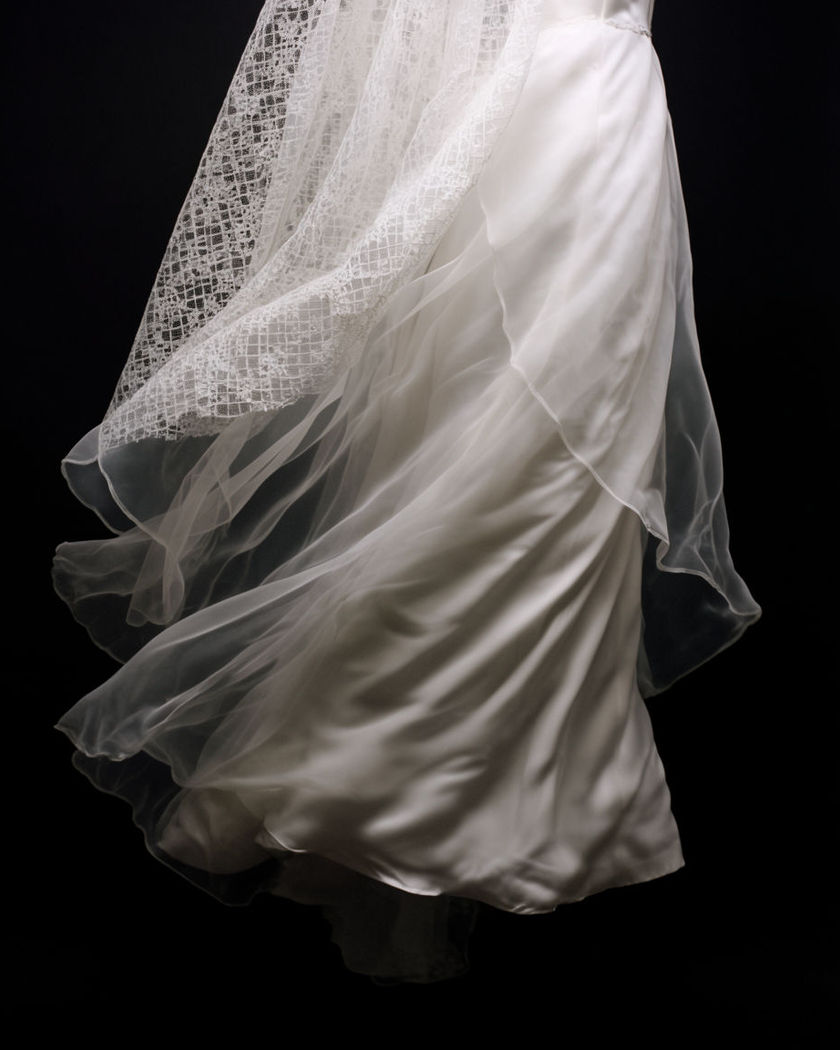 A flowing white bridal gown and sheer veil catching soft light, fabric mid-transformation against a black ground