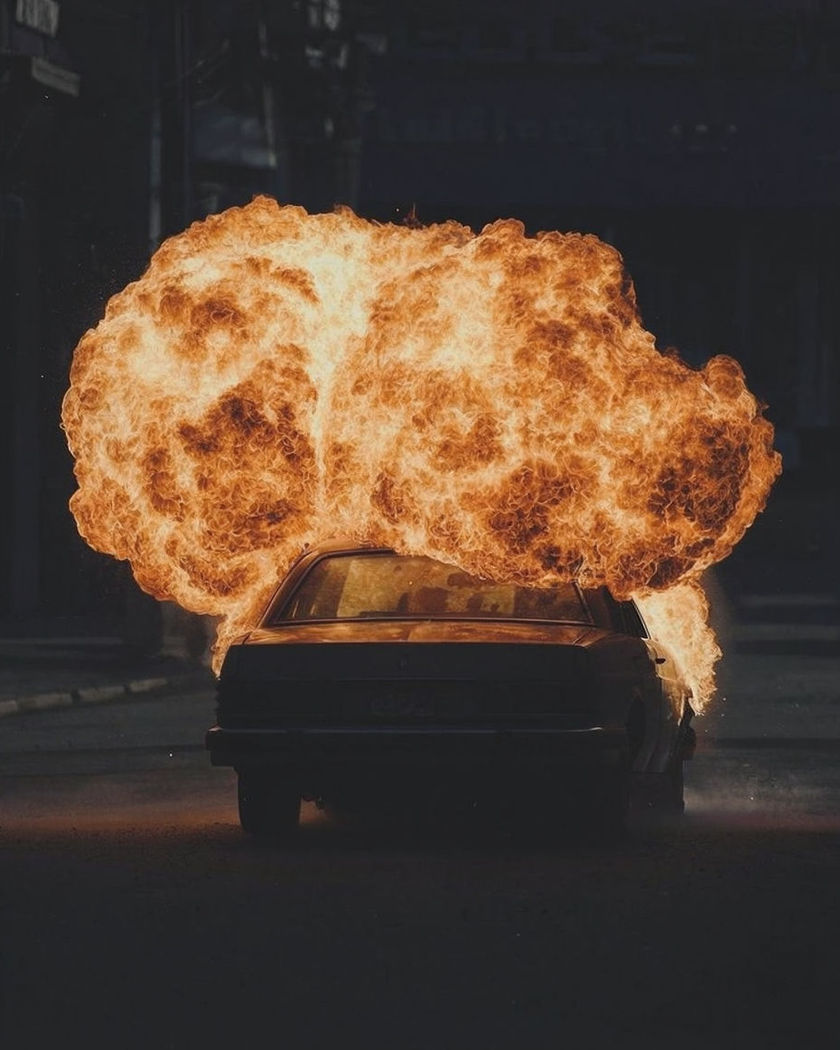 A car mid-detonation, a rolling orange fireball lifting off the chassis against a dark street