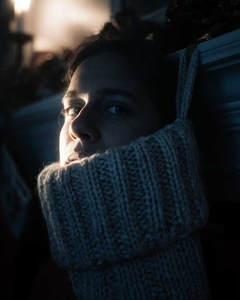 A face peeking out of the top of an oversized knitted Christmas stocking, warm mantel lights glowing behind