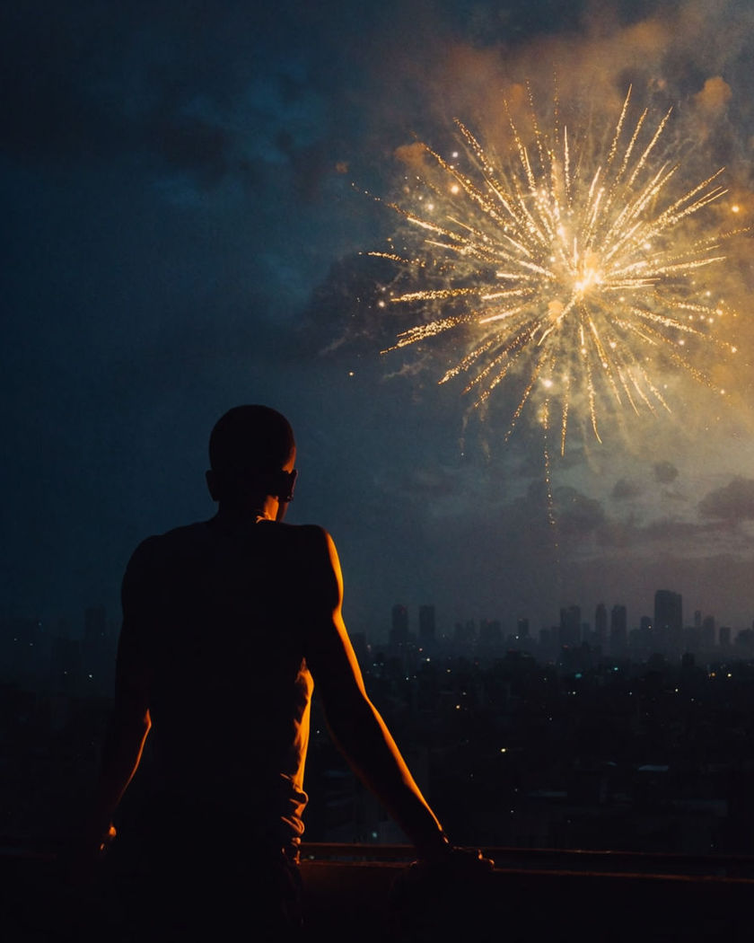 A silhouette on a rooftop as a burst of gold fireworks blooms over a generic invented city skyline