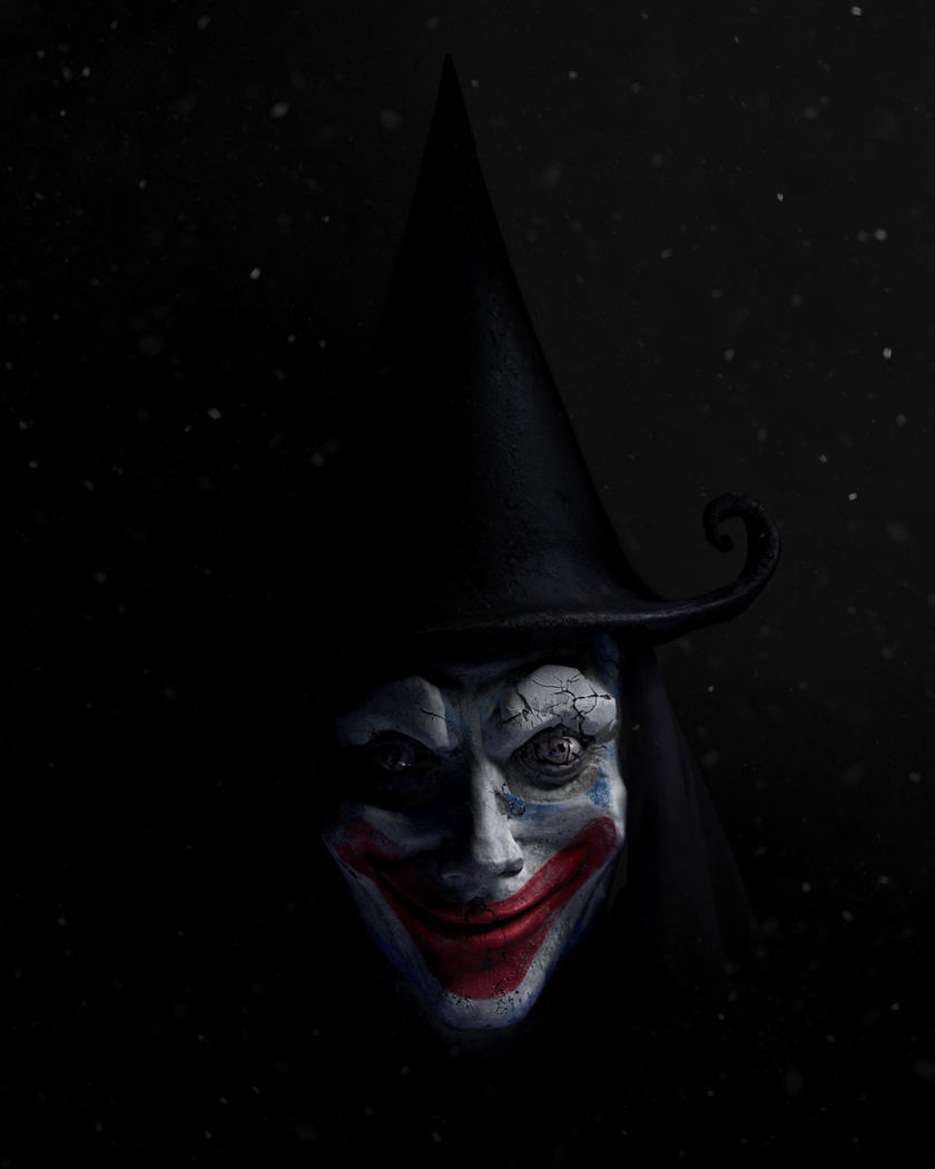 A pale greasepainted horror clown face with a cracked red grin emerging from deep shadow under a single dim light