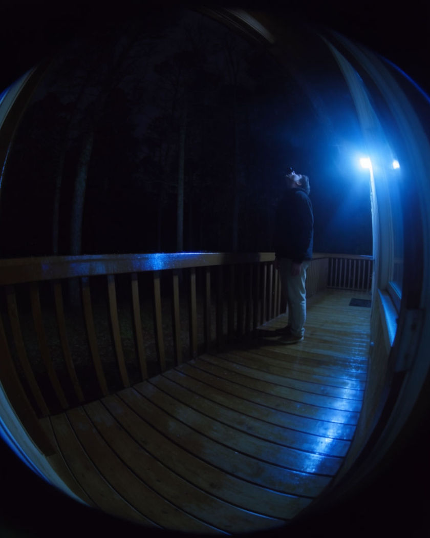 A wide fisheye doorbell camera view at night showing a figure pausing on a porch and looking up at the lens