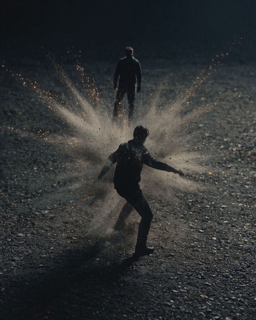 A silhouette bursting outward into a violent radial cloud of ash and glowing particles against a dark ground